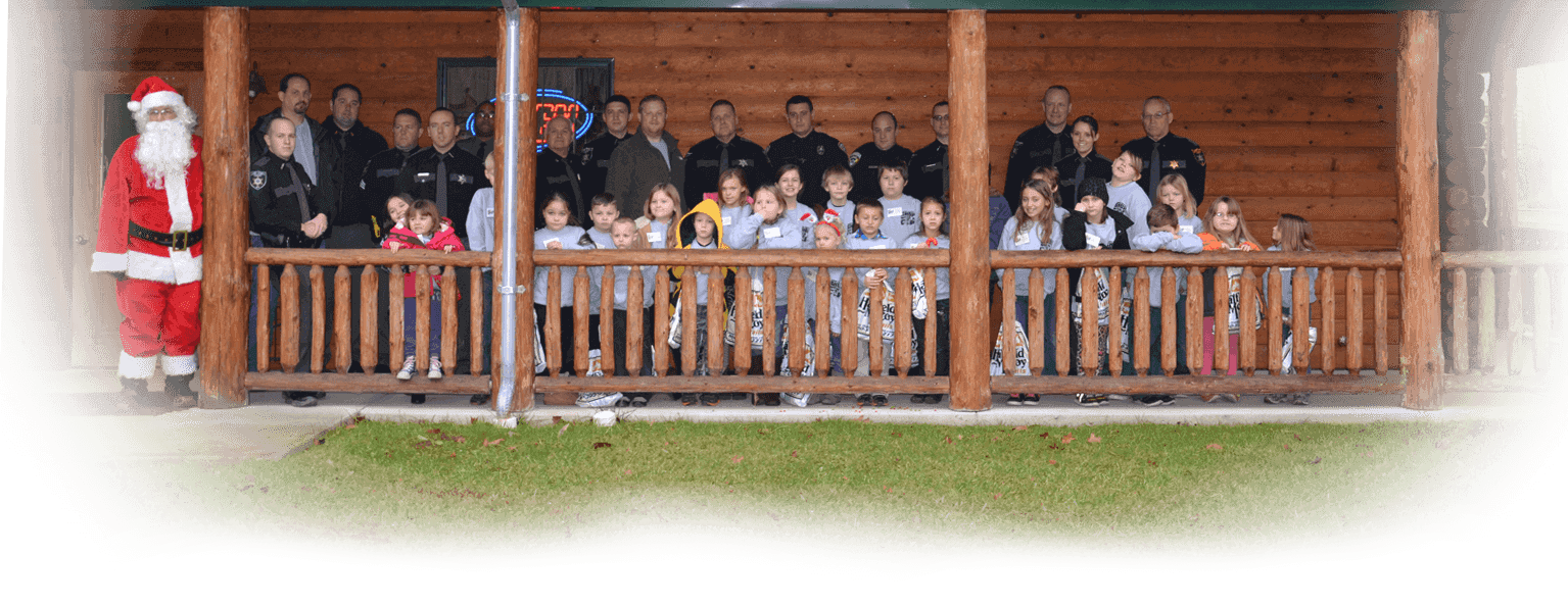 Image of kids and officers standing in front of a log building with santa in frame. The children are all smiling.